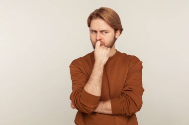 Düşünceli, kafası karışmış, sakallı, eşofman üstü giyen, ikilemi düşünüp karar veren ya da zor bir karar veren düşünceli bir adamın portresi. Gri arka planda kapalı stüdyo çekimi