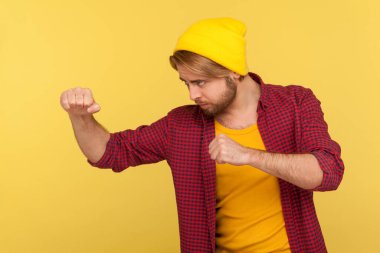 Şapkalı kararlı hippi sakallı adam, kareli gömlekli boks yapmaya hazır yumruklar tutan kendinden emin ifadeyle yumruk atan, savaşçı ruhlu. Stüdyo görüntüsü sarı, yan görünüm