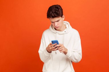 Kapüşonlu kapüşonlu tişörtlü, elinde cep telefonu olan, web sayfalarını kaydıran, internetten yemek sipariş eden ciddi konsantre bir genç. Turuncu arka planda kapalı stüdyo çekimleri
