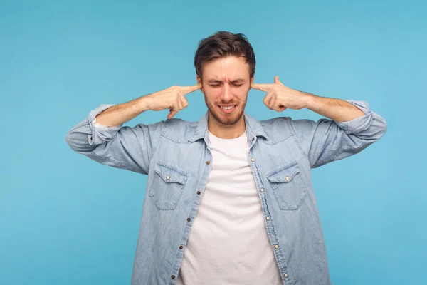 Don\'t want to listen! Portrait of man in worker denim shirt closing ears and grimacing displeased, irritated by noise, difficult to hear annoying sound. indoor studio shot isolated on blue background