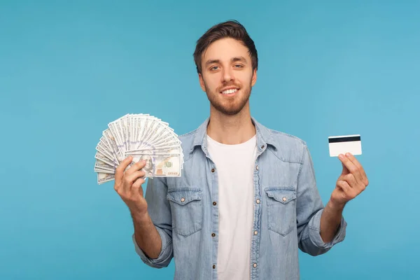 送金だ 労働者のデニムシャツの幸せなハンサムな男の肖像画は おもちゃの笑顔でカメラを見て ドル紙幣やクレジットカードを示しています 青い背景に隔離された屋内スタジオで — ストック写真