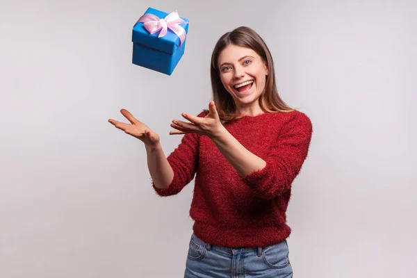 可爱快乐的黑发女孩穿着毛茸茸的毛衣 抛掷并抓起礼品盒 无忧无虑地笑着 享受节日礼物的形象 在灰色背景下隔离的室内拍摄 — 图库照片