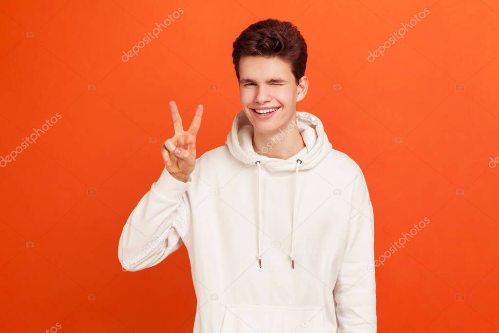 Happy positive teenager in stylish sweatshirt showing v sign to camera, winking and smiling, young man wishing good luck, peace. Indoor studio shot isolated on orange background