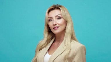 İş stili ceketli, neşeli, sarışın bir kadın göz kırpıyor, gülümsüyor, cilveli, çekici bir bayanla flört ediyor. Mavi arka planda kapalı stüdyo çekimi