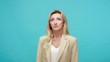 İş ceketi giymiş üzgün yetişkin bir kadın parmaklarını alnına yakın tutarak ezik işaretini alaycı bir şekilde gösteriyor. Kriz yüzünden mutsuz. Mavi arka planda kapalı stüdyo çekimi