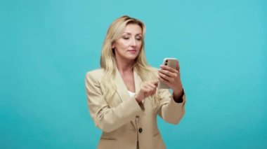 Sarı saçlı yetişkin iş kadını akıllı telefon kullanıyor, web sayfalarını kaydırıyor ve sosyal ağda sohbet ediyor, şaşırmış bir suratla zum yapıyor. Mavi arka planda kapalı stüdyo çekimi