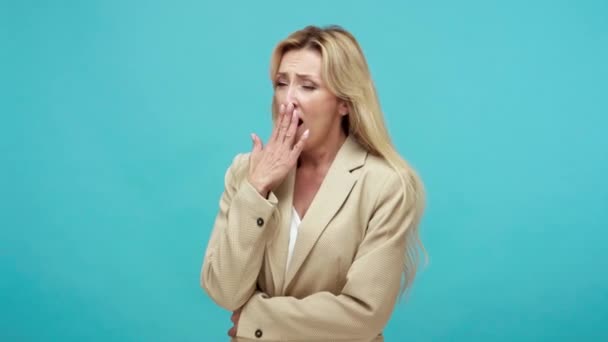 ビジネスジャケットで疲れた眠い大人のブロンドの女性は退屈な話を聞いて手で口を閉じてあくびを 青い背景に隔離された屋内スタジオショット — ストック動画
