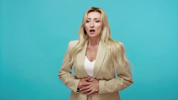 ビジネススタイルのジャケットで大人のブロンドの女性は 胃の痛み 消化不良 痛みの期間 流産を感じ痛みで嘆き悲しむ 青い背景に隔離された屋内スタジオショット — ストック動画