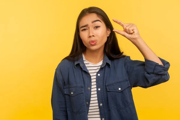 Too small! Portrait of displeased girl in denim shirt showing a little bit gesture, dissatisfied with size measuring low scale, minimum inch or centimeter. studio shot isolated on yellow background