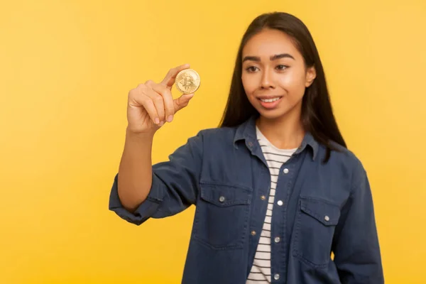 Cryptocurrency btc coin. Portrait of rich happy brunette girl in denim shirt smiling and looking at golden bitcoin, electronic money, digital currency. indoor studio shot isolated on yellow background