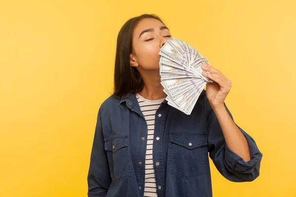 Greedy for money. Portrait of avaricious rich girl in denim shirt smelling earned dollar banknotes, enjoying success and big profit, wealthy life. indoor studio shot isolated on yellow background