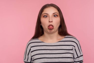 Portrait of funny dumb amusing woman in striped sweatshirt standing with crossed eyes, showing tongue out and making silly brainless facial expression. indoor studio shot isolated on pink background 