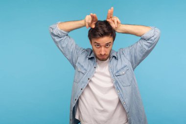 Kot pantolon giymiş saldırgan bir adamın portresi boğa boynuzu jesti ile kameraya tehditkar bir şekilde bakıyor, saldırmakla tehdit ediyor, çatışmayı kışkırtıyor. Stüdyo çekimi mavi arkaplanda izole edildi 