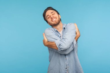 Kendimi seviyorum! Kendini kucaklayan, egosu, zevki ve özgüveni ile gülümseyen işçi kot gömleği giymiş bencil narsist bir adamın portresi. Stüdyo çekimi mavi arkaplanda izole edildi 