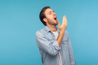 Dinlenmem lazım! Çalışan kot gömlekli yorgun bir adamın portresi esniyor ve uyuma hayali kuruyor, uykulu hissediyor, iş yapmak için yetersiz, tembellik ve enerji eksikliği. Stüdyo çekimi mavi arkaplanda izole edildi 