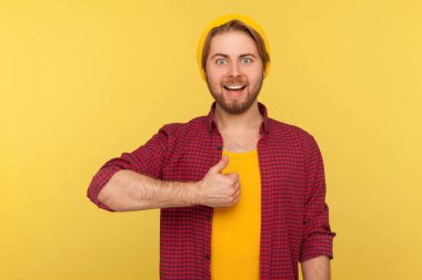 Yani, aferin! Bereli şapkalı, damalı tişörtlü, mutlu, sakallı hippi adam. Başparmak kaldırıyor, onay veriyor, başarı işareti yapıyor. Sarı arka planda izole edilmiş kapalı stüdyo çekimi 