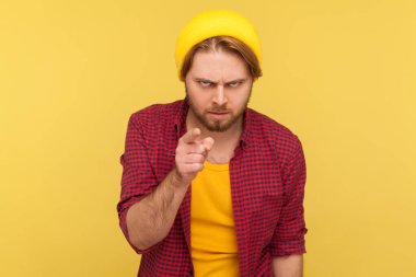 Hey sen, dikkatli ol! Katı hippi sakallı, bereli ve damalı tişörtlü adam kameraya bakıyor, kızgın ve uyarıyor, otoriter yüzüyle alarm veriyor. Stüdyo çekimi sarı arkaplanda izole edildi 