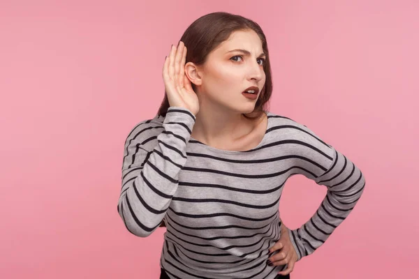 What? I can\'t hear you! Portrait of confused woman in striped sweatshirt keeping hand near ear to listen better, having hearing problems, difficult to understand. indoor studio shot, pink background