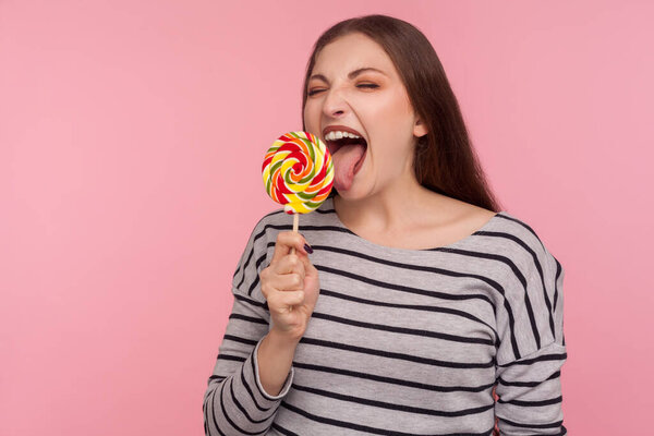 Portrait of woman in striped sweatshirt licking lollipop, tasting sweet round rainbow candy with crazy expression, enjoying delicious flavor dessert. indoor studio shot isolated on pink background 