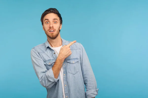 Wow, look at ads here! Portrait of surprised shocked man in denim shirt pointing aside, showing blank copy space for idea presentation, commercial text. indoor studio shot isolated on blue background