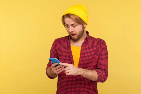 Amazed hipster guy in beanie hat and checkered shirt looking surprised at cell phone, reading shocking message, chatting in social network, astonished by mobile app. indoor studio shot isolated