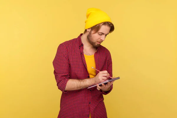 ビーニー帽子のヒップスタートレンディーな男は 創造的なアイデアに触発され 熱心にノートブックにメモを取り ビジネスプランにメモを取ることを思い出させる書きを書く 黄色で隔絶された屋内スタジオで — ストック写真