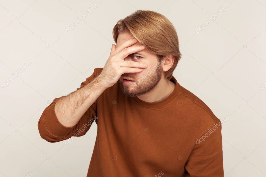 Portrait of nosy inquisitive bearded man peeking covering face and eyes with hand, looking through fingers with curious expression, spying or shy to watch. studio shot isolated on gray background 