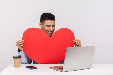 Yakışıklı, mutlu adam masada oturuyor, kırmızı kalbi tutuyor ve bilgisayardan video görüşmesi yapıyor, gülümsüyor, romantik duygular ifade ediyor, internetten biriyle çıkıyor. Beyaz arka planda izole edilmiş kapalı stüdyo çekimi 