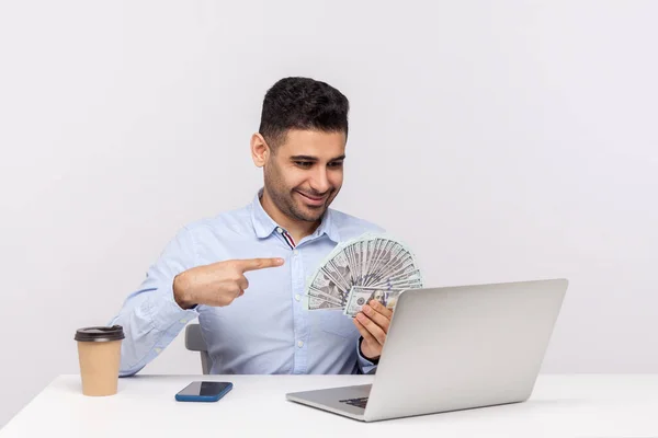 Successful happy elegant businessman sitting in office workplace pointing money dollars and smiling at laptop screen, having online conversation, boasting financial profit. indoor studio shot isolated