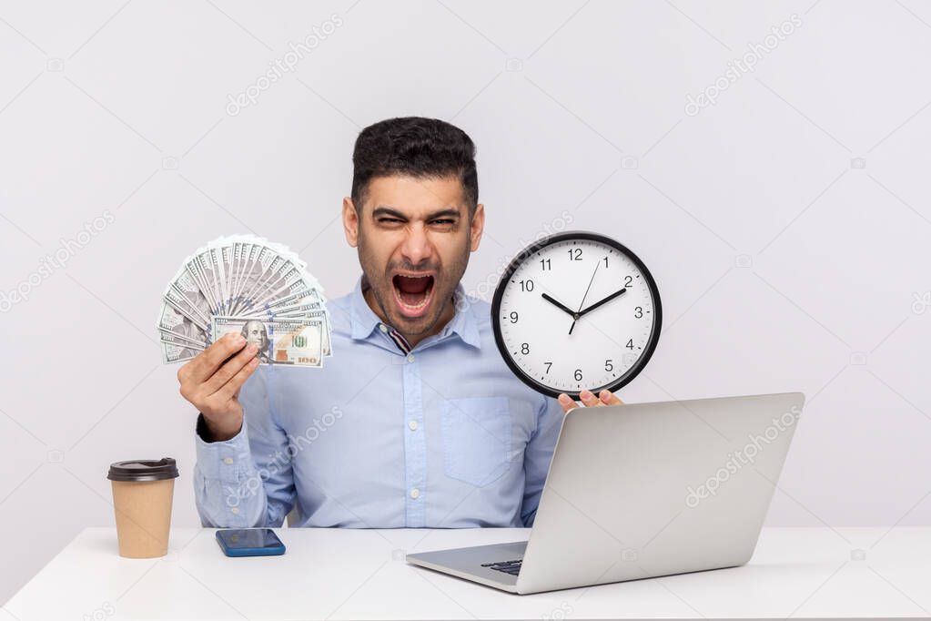 Time is money! Aggressive businessman sitting in office workplace, holding big clock and dollar banknotes, shouting with anger, looking furious crazy. indoor studio shot isolated on white background 