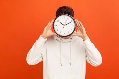 Don't waist your time! Man in casual white sweatshirt holding wall clock hiding his face, time management, schedule and meeting appointment. Indoor studio shot isolated on orange background  clipart