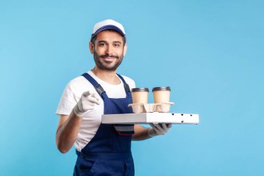 Teslimat servisi. Tulum giymiş, elinde kahve, pizza kutusu olan mutlu bir kurye güvenlik eldiveni takıp içki ikram ediyor, kamerayı işaret edip seni seçiyor. Kapalı stüdyo çekimi, izole.