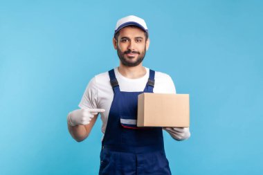Kargo taşımacılığı, teslimat servisi. Üniformalı dost canlısı işçi karton kutuyu işaret ediyor ve kameraya gülümsüyor. Paketi taşıyan bir kurye. Çevrimiçi ekspres nakliye, taşıma hizmeti. izole