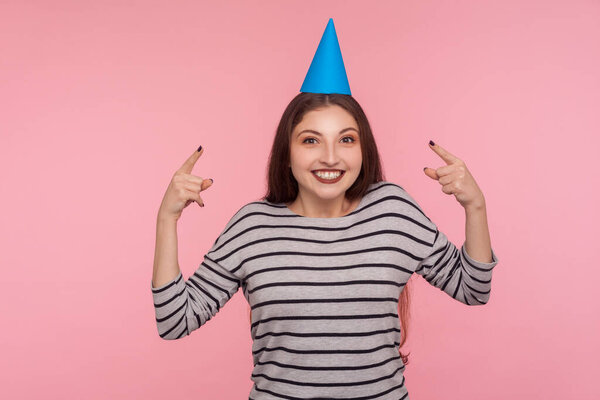 Happy birthday to me! Portrait of optimistic pretty woman pointing at party cone hat and looking at camera with toothy smile, rejoicing holiday celebration. studio shot isolated on pink background