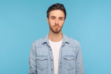 Şık saç stili ve kıllı, işçi kot gömleği giyen ve kameraya dikkatlice odaklanmış ifadesiyle bakan ciddi yakışıklı bir gencin portresi. Stüdyo çekimi mavi arkaplanda izole edildi