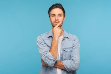 Hmm, düşünelim! İşçi kot gömleği giymiş düşünceli yakışıklı bir adamın portresi plan düşünürken çenesine dokunuyor, zor seçimler hakkında şüpheleri var, emin değilim. Stüdyo çekimi mavi arkaplanda izole edildi