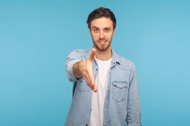 Merhaba, tanıştığımıza memnun oldum! Dost canlısı gülümseyen yakışıklı adam portresi işçi kot gömleği içinde uzanan, iş görüşmesinde el sıkışma teklif eden. Mavi arka planda kapalı stüdyo çekimleri