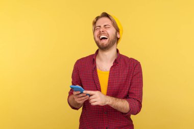 Şapkalı ve damalı gömlekli neşeli hippi çocuk cep telefonu kullanıyor, sosyal ağda mesaj okuduktan sonra yüksek sesle gülüyor, internette komik şakalar yapıyor. Stüdyo çekimi sarı arkaplanda izole edildi