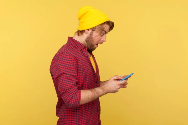 サイドビューは 携帯電話で驚きを見て ショッキングなメッセージを読み ソーシャルネットワークでチャットし モバイルアプリに驚いているヒップスタービーニー帽子とシャツの男を驚かせた 屋内スタジオでの撮影は — ストック写真