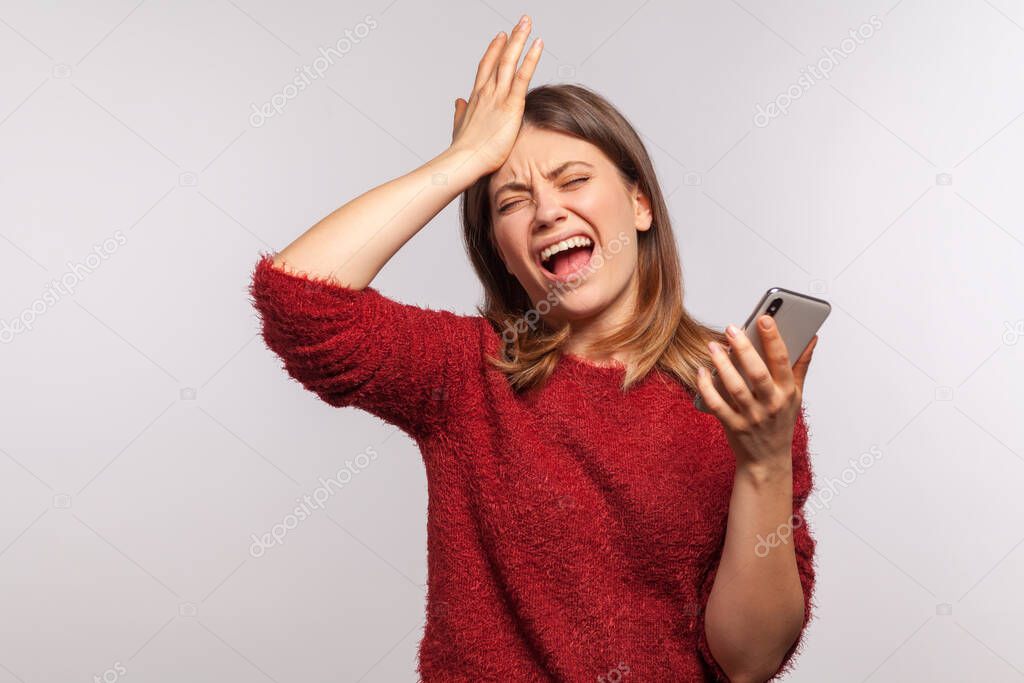 Portrait of upset depressed woman in shaggy sweater standing with facepalm gesture and holding mobile phone, expressing sorrow regret, blaming herself. indoor studio shot isolated on gray background