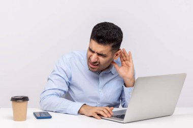 Kafası karışmış işadamı işyerinde oturuyor, dizüstü bilgisayara eğiliyor ve el ele tutuşup daha iyi duymaya çalışıyor, sessiz sesleri dinliyor. Beyaz arka planda izole edilmiş kapalı stüdyo çekimi