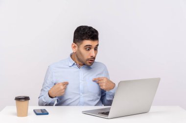 Gerçekten, ben mi? Şaşırtıcı iş adamı ofis işyerinde oturuyor, kendini gösteriyor ve şok olmuş bir ifadeyle dizüstü bilgisayarına bakıyor, video konuşmasında konuşuyor. Beyaz arka planda izole edilmiş kapalı stüdyo çekimi