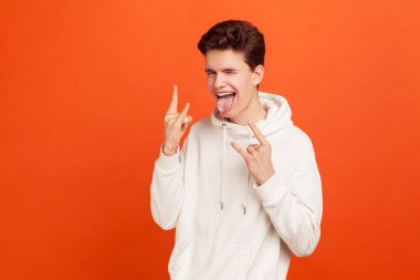 Sıradan beyaz kapüşonlu genç adam rock 'n roll işareti el hareketi yapıyor ve göz kırpıyor, dil gösteriyor. Aktif yaşam tarzı, performans. Turuncu arka planda kapalı stüdyo çekimleri