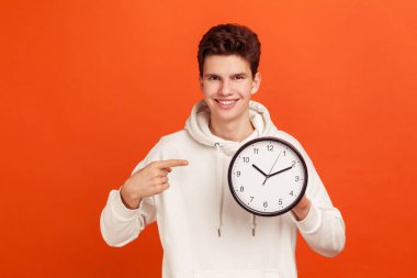 Cheerful confident young man in casual sweatshirt with hood pointing finger on clock in his arm, time to act, start-up. Indoor studio shot isolated on orange background clipart