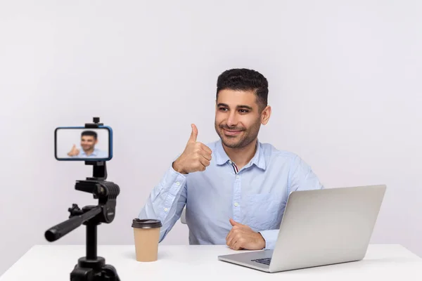 Online webinar, distant training. Elegant successful businessman sitting at workplace, showing thumbs up to mobile phone while recording course, doing blog. studio shot isolated on white background