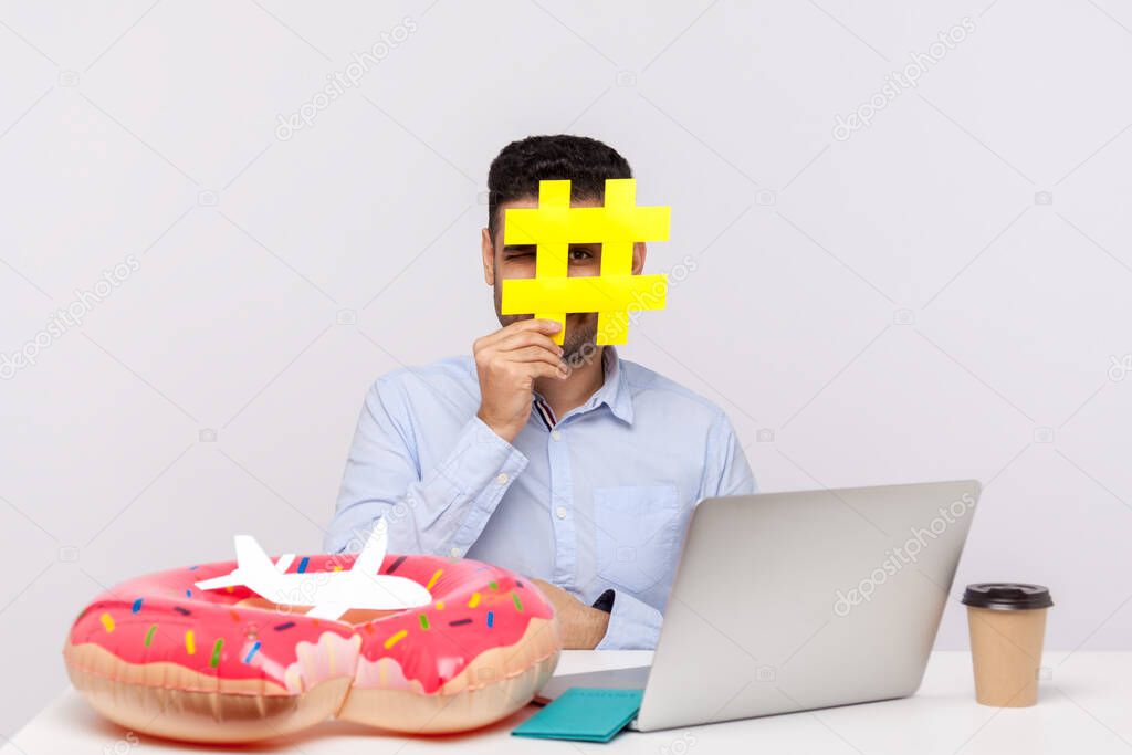 Time to rest! Man looking through hashtag symbol, sitting in office workplace with rubber ring passport and paper airplane on desk, holding hash sign of viral web topic about relax travel. studio shot