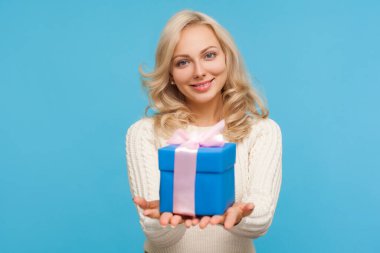 Cute positive blond woman in knitted sweater holding in hands blue giftbox with ribbon bow, making present, surprise. Indoor studio shot isolated on blue background clipart