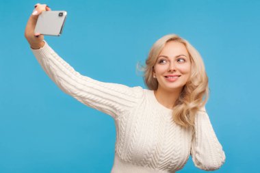 Örgü kazaklı şık bir kadın elinde akıllı telefon, sosyal ağ için selfie çeken bir kadın blogcu. Mavi arka planda kapalı stüdyo çekimi