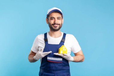 Mutlu tamirci portresi, iş kıyafetleri içinde nakliyeci ve kağıt ev gösteren eldivenler. Ev tamiri, bakım ve taşıma hizmetleri, taşınma konusunda yardım. Stüdyo çekimi mavi arkaplanda izole edildi