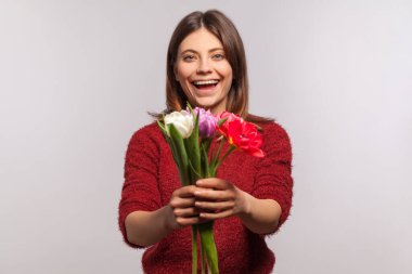 Kameraya çiçek buketi verip heyecanla gülümseyen neşeli esmer kızın portresi. Bahar tatilini kutlarım, 8 Mart Kadınlar Günü. Gri arka planda kapalı stüdyo çekimi